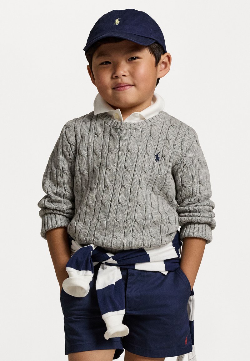 Šedý sveter s pleteným vzorom, biela košeľa s golierom, námornícke šortky a námornícka čiapka s logom. Šortky sú stiahnuté bielou a námorníckou pruhovanou látkou.