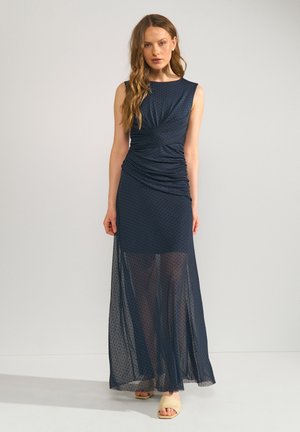 Femme portant une robe sans manches bleu marine à pois froncée avec superposition de jupe transparente et sandales beiges à bout ouvert, debout devant un fond uni.