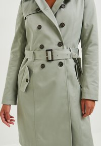 Světle zelený trenčkot s dvojitým zapínáním vpředu, velkými knoflíky a opaskem v pase. Obsahuje boční kapsu a hladkou texturu.