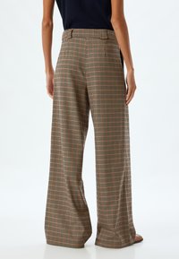 Geruite wijde broek in beige, bruin en rode tonen. Heeft een hoge taille, plooien en een subtiele textuur. Achteraanzicht weergegeven.