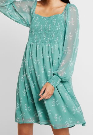 Frau trägt ein türkisfarbenes Blumenkleid mit transparenten langen Ärmeln, gerafftem Oberteil und einem fließenden Rock, hält den Saum mit einer Hand.