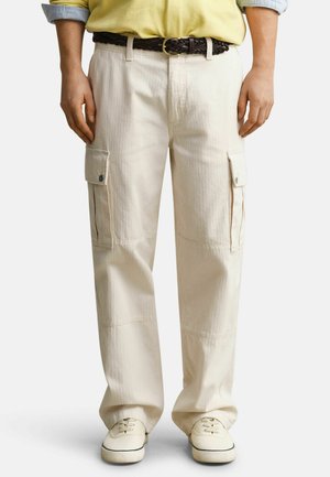 Persoon draagt beige cargobroek met zijzakken, een gevlochten bruine riem, geel shirt en witte sneakers tegen een effen achtergrond.