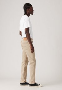 Beige jeans met een rechte snit, voorzien van zichtbare stiksels, een leren label op de tailleband en zwarte canvas sneakers. Witte t-shirt.