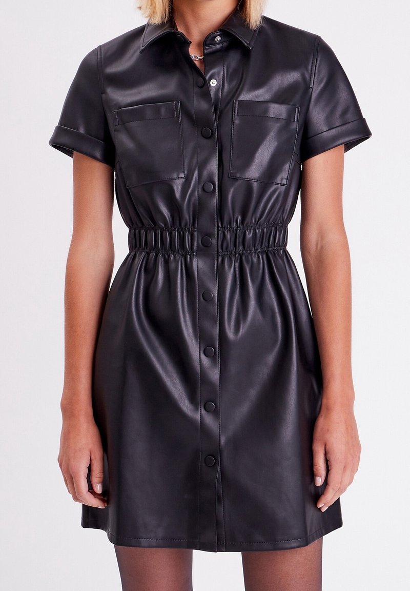 Robe chemise en simili cuir noir avec manches courtes, fermeture boutonnée, deux poches poitrine et taille élastiquée pour une silhouette ajustée.