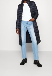 Lång svart puffjacka med hög krage, kombinerad med ljusblå jeans och svarta ankelstövlar. Slät, vadderad textur, elegant design.