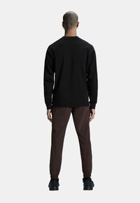 Camicia nera a maniche lunghe con texture liscia, abbinata a pantaloni marrone scuro con polsini elasticizzati e sneakers nere. Vista da dietro.