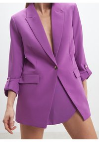 Lila kavaj i mjukt tyg, med djup V-ringning, långa ärmar med uppvikta manschetter och framfickor med klaff. Guldknappar som accent.