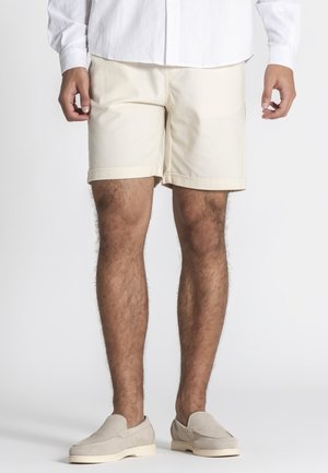 Mand iført beige shorts, lysebeige slip-on sko og en hvid langærmet skjorte, stående på en ensartet lys baggrund.