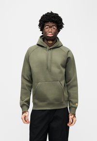 Πράσινο φούτερ με κουκούλα, μπροστινή τσέπη, ριμπ μανσέτες και μικρή διακριτική στάμπα στο μανίκι· μαλακό, υφασμάτινο με υφή.