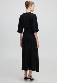 Abito lungo nero con una texture a maglia, dotato di maniche oversize, una vita aderente e pannelli trasparenti all'orlo. La vista posteriore evidenzia i dettagli dello scollo.