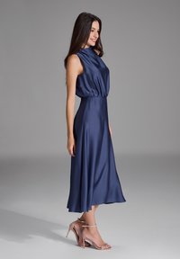 Robe en satin bleu marine avec une taille cintrée et une jupe évasée, présentant un décolleté haut et une coupe sans manches. Associée à des talons à brides transparentes.