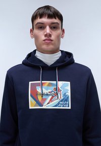 Námorníckou mikinu s prednou grafikou, ktorá zobrazuje lyžiarsky dizajn, farebné geometrické tvary a kontrastné šnúrky. Mäkka textúra, voľný strih.