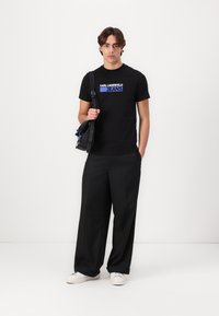 Juodas medvilninis marškinėlis su mėlynu logotipu "KARL LAGERFELD JEANS." Suderintas su plačiomis juodomis kelnėmis ir baltais sportbačiais. Per petį nešiojama kuprinė.