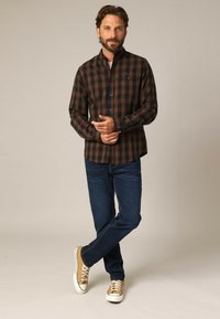 Bruine en zwarte geruite button-up shirt met een kraag, gecombineerd met donkere spijkerbroek en lichtbruine sneakers. Heeft een borstzak en lange mouwen.