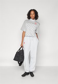 Top scurt cu mâneci scurte în carouri gri, cu un logo roșu, pantaloni albi de trening și o geantă de purtat neagră. Încălțăminte sport neagră cu prindere texturată.