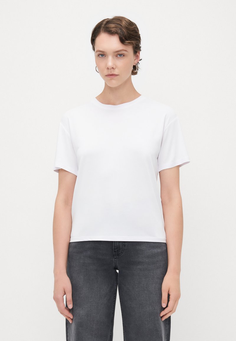 T-shirt bianca a maniche corte con colletto rotondo, caratterizzata da una texture liscia e una vestibilità rilassata, abbinata a jeans grigio scuro.