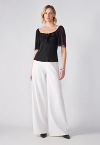 Top negru cu maneci scurte bufante si decolteu fronsat, combinat cu pantaloni albi cu picioare largi. Modelul stă pe un fundal simplu.
