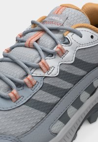 Gros plan sur une basket Merrell grise avec un tissu texturé, des accents orange, des lacets croisés et une semelle au design robuste.