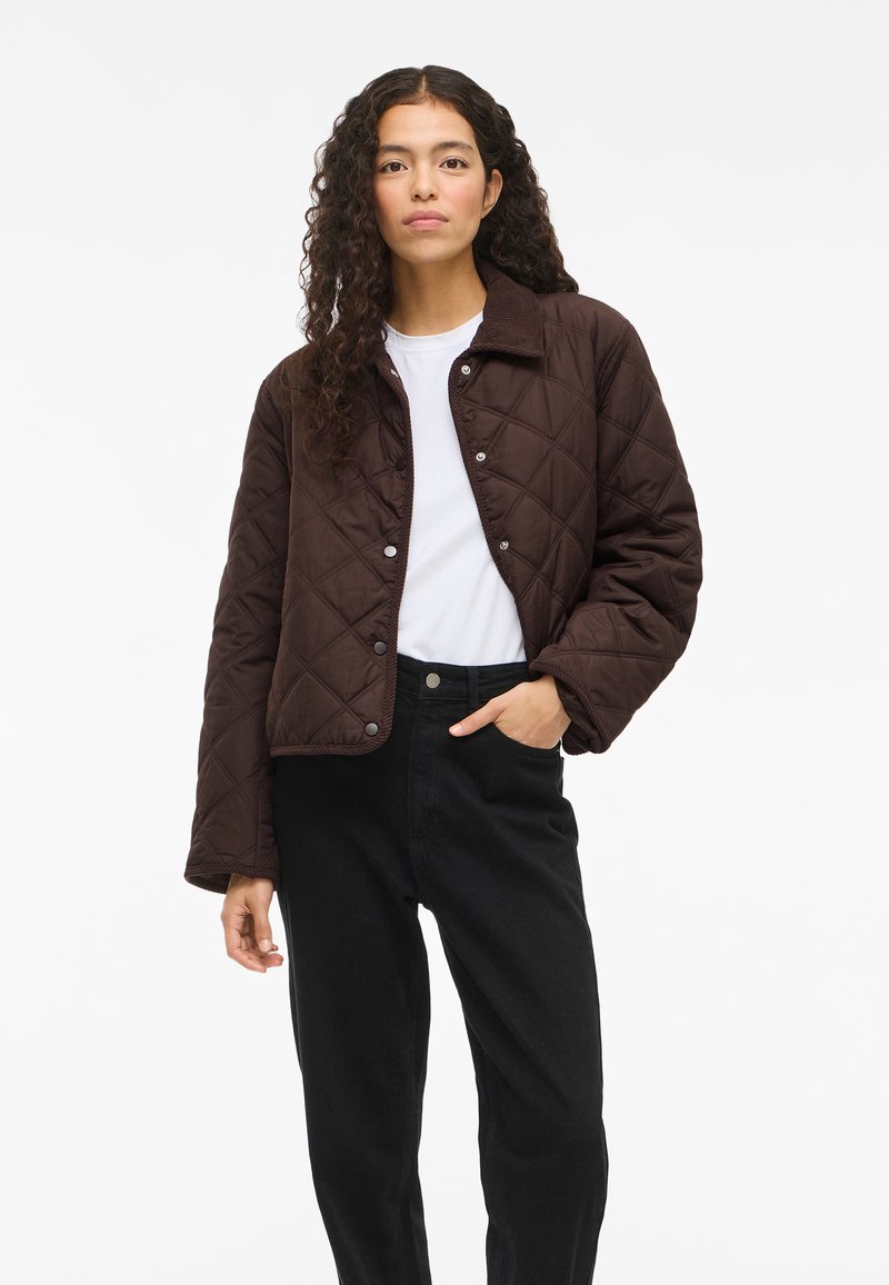 Mulher com cabelo encaracolado escuro, vestindo um casaco acolchoado castanho sobre uma camisa branca e calças pretas, em pé com uma mão no bolso.
