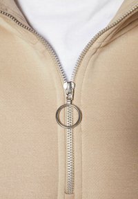 Giacca beige con zip, dal texture morbido, con una cerniera argentata e un tirante circolare. Sotto c'è una camicia bianca.