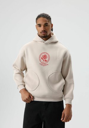 Beige hoodie met een voorkantgrafiek van een roos, grote zijzakken, geribbelde manchetten en een capuchon met trekkoord. Draagt donkere jeans.