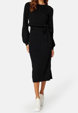 Femme portant une robe noire en maille côtelée à manches longues, taille nouée, et baskets blanches, debout devant un fond clair et uni.