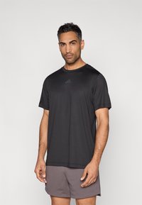 Μαύρο αθλητικό t-shirt με κοντά μανίκια, με αντανακλαστικό λογότυπο στο στήθος. Συνδυάζεται με ανοιχτό γκρι σορτς, με απαλή υφή υφάσματος.