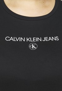 Černé přiléhavé tričko s kulatým výstřihem, které má bílé nápisy "CALVIN KLEIN JEANS" a kruhové logo "CK". Hladká textilie.