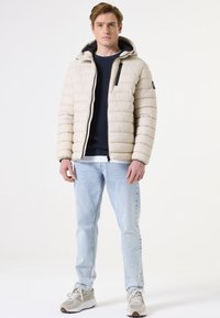Beige gepolsterte Jacke mit Kapuze, Frontreißverschluss und schwarzen Akzenten. Über einem marineblauen Pullover getragen, kombiniert mit hellblauen Jeans und Sneakers.