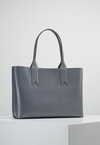 Szara skórzana torba tote o wyraźnej konstrukcji, z dwiema płaskimi uchwytami i trójkątnymi akcentami u podstawy uchwytów.
