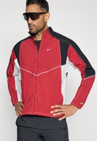 Sarkans, melns un balts uzjūgšanas jakas, izgatavotas no viegla materiāla; ar augstu apkakli, sānu kabatām un kontrastējošām šuvēm. Redzams Nike logo.