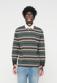 Langermet polo skjorte i mørkegrønn, brun, og hvite striper. Har en kontrasterende hvit krage og en knapp nedover halsen. Kombinert med lyseblå jeans.