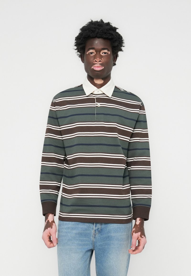 Langermet polo skjorte i mørkegrønn, brun, og hvite striper. Har en kontrasterende hvit krage og en knapp nedover halsen. Kombinert med lyseblå jeans.