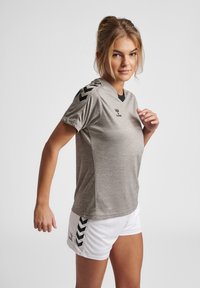 Grå atletisk skjorte med sorte accenter, kortærmer og V-hals design, parret med hvide shorts med sort chevron mønster.