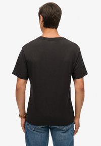 Sort T-shirt med korte ærmer, rund halsudskæring, glat tekstur og løs pasform, båret med blå jeans. Set fra bagsiden.