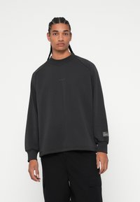 Tumši pelēks džemperis, izgatavots no mīkstas auduma, ar raglanu piedurknēm, ribotu apkakli un diskretu zīmolu uz priekšpuses un piedurknes.