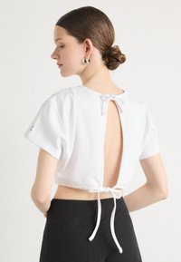 Λευκό crop top από μαλακό ύφασμα, με κοντά μανίκια, κοπή στην πλάτη και κούμπωμα με κορδόνι. Μινιμαλιστικός σχεδιασμός χωρίς σχέδια ή διακοσμητικά στοιχεία.