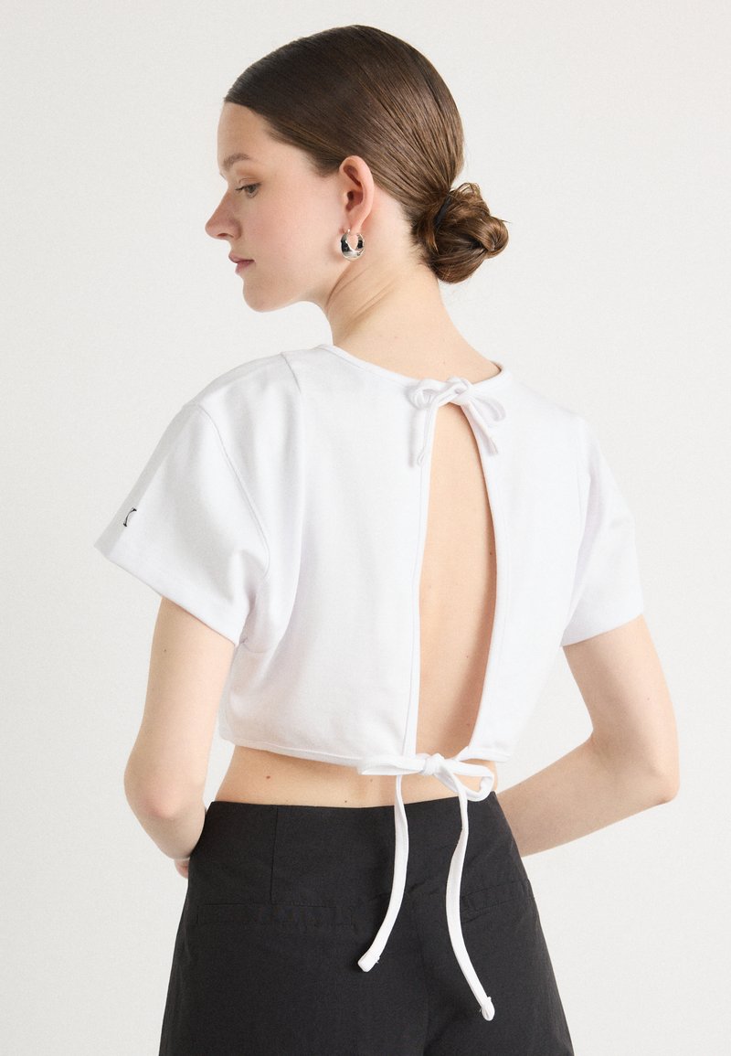 Λευκό crop top από μαλακό ύφασμα, με κοντά μανίκια, κοπή στην πλάτη και κούμπωμα με κορδόνι. Μινιμαλιστικός σχεδιασμός χωρίς σχέδια ή διακοσμητικά στοιχεία.