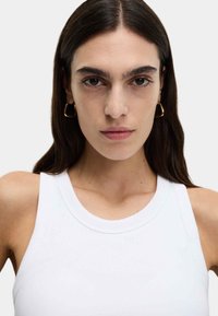 Weißes geripptes Tanktop mit rundem Ausschnitt und breiten Trägern, kombiniert mit goldenen Creolen, vor neutralem Hintergrund.
