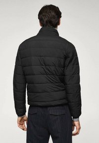 Veste matelassée noire avec un col montant, présentant des coutures horizontales et des poignets élastiques. Le tissu a une texture lisse.