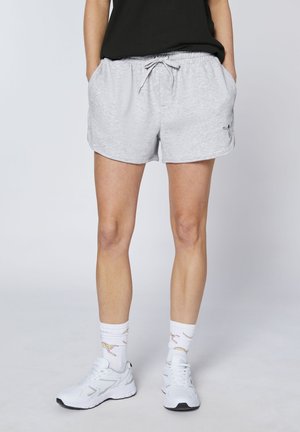 Person wearing light gray drawstring shorts, black top, white socks with cheetah print, and white athletic shoes, standing with hands in pockets.