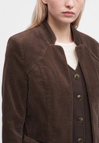 Femme portant une veste en velours marron sur un gilet marron foncé boutonné et un top en maille blanche.