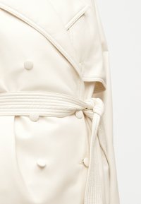 Καμπαρντίνα από δέρμα εκρου με κουμπιά στο ίδιο χρώμα και ζώνη με ραφές δεμένη σε κόμπο στη μέση.