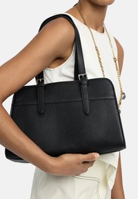 Osoba w bezrękawniku białym i złotym łańcuszku, trzymająca czarną torebkę ze strukturalnej skóry z dwoma ramiączkami na ramię.