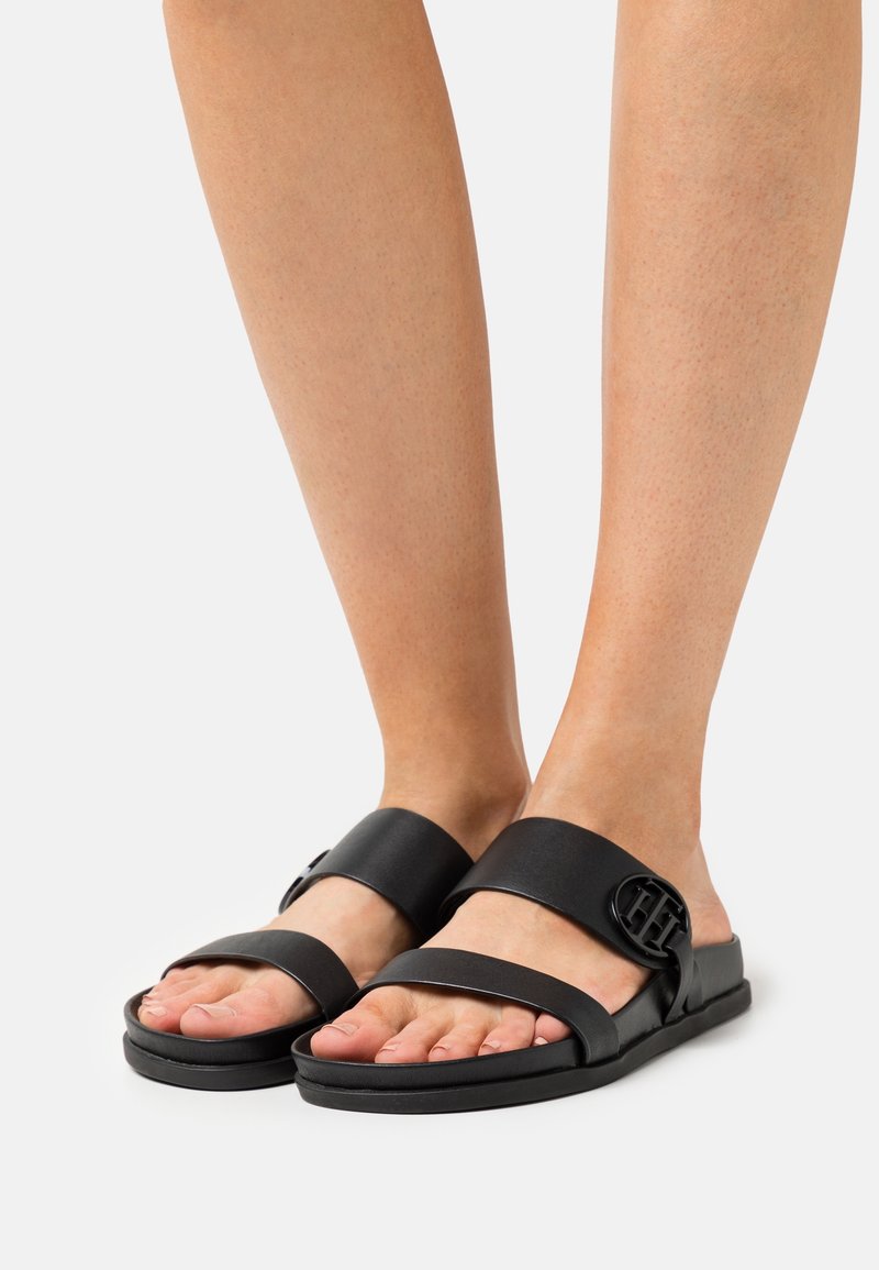 Pieds chaussés de sandales plates noires avec deux brides et un détail de boucle ronde sur la bride supérieure, sur fond blanc.