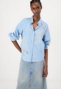Camisa azul clara com riscas, com um design floral bordado, frente com botões e mangas arregaçadas, combinada com uma saia de denim.