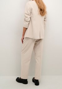 Blazer beige sur mesure avec épaules structurées et manches trois-quarts, assorti d'un pantalon ample assorti ; chaussures à plateforme noires.