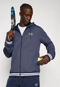 Sweatshirt com fecho éclair da marinha, com punhos listrados em branco, tecido texturizado e logótipo EA7. A pessoa segura uma raquete de ténis com uma mão e uma bola de ténis com a outra.