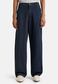Persoon in donkerblauwe wijde jeans en witte loafers, met ontspannen handen langs de zijkanten, op een effen lichte achtergrond.