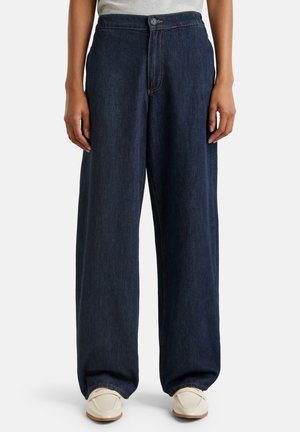 Personne portant un jean large bleu foncé et des mocassins blancs, les mains détendues sur les côtés, sur un fond clair uni.